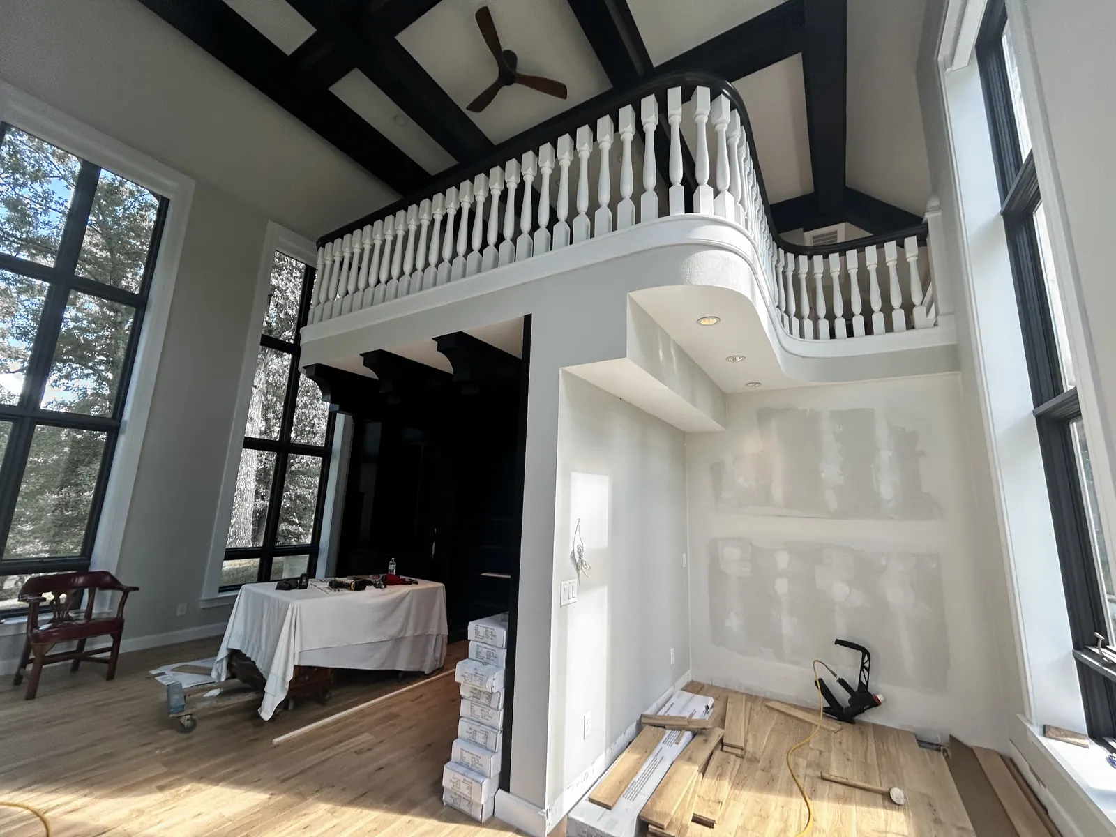A two-story room during construction: boxes of flooring, a pneumatic flooring nailer and loose boards on a partly installed floor, under a curved balcony railing